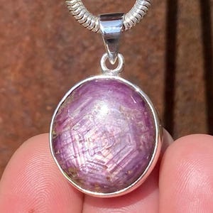 May include: A round, purple gemstone pendant with a silver bezel and bail. The gemstone has a star-like pattern within it. The pendant hangs from a silver chain. The background is a blurred, reddish-brown colour.