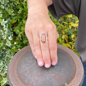 May include: A silver ring with an oval-shaped amber stone set in a silver bezel. The ring is on a person's finger.