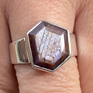 May include: A silver ring with a large, faceted, hexagonal gemstone. The gemstone is a deep burgundy color with internal light-colored inclusions. The ring band is wide and smooth, with a polished finish. The ring is worn on a finger.