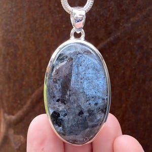 May include: Oval labradorite pendant in a silver setting. The gemstone shows blue, grey, and black colours. The pendant is suspended from a silver chain. The background is a blurred brown surface.