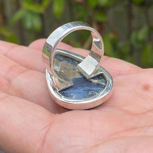 May include: A silver ring with a teardrop-shaped, blue and brown stone. The ring's band is thick and polished, with a simple design. The stone has a marbled appearance, with swirls of blue, brown, and white. The ring is set on a hand.