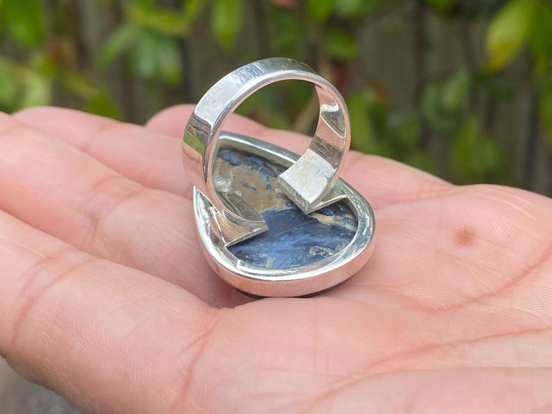 May include: A silver ring with a teardrop-shaped, blue and brown stone. The ring's band is thick and polished, with a simple design. The stone has a marbled appearance, with swirls of blue, brown, and white. The ring is set on a hand.