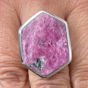 May include: A silver ring with a large, pink, hexagonal, faceted gemstone. The gemstone has a rough, natural texture.