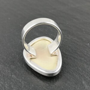 K&ouml;nnte beinhalten: Ein silberner Ring mit einem wei&szlig;en ovalen Edelstein, der in der Mitte eingesetzt ist. Der Ring wird von hinten gezeigt, wodurch die Fassung und die Ringschiene sichtbar werden.