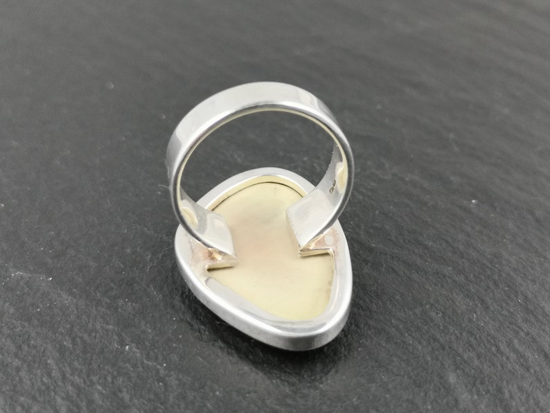 K&ouml;nnte beinhalten: Ein silberner Ring mit einem wei&szlig;en ovalen Edelstein, der in der Mitte eingesetzt ist. Der Ring wird von hinten gezeigt, wodurch die Fassung und die Ringschiene sichtbar werden.