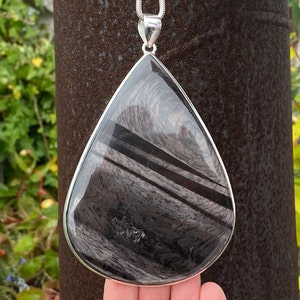 Pode incluir: Um pingente de prata com uma grande pedra de obsidiana preta em forma de lágrima. A pedra tem um padrão de redemoinho e estriado.