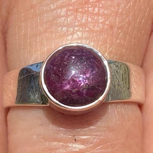 Könnte beinhalten: Ein Silberring mit einem großen, runden, tiefvioletten Edelstein. Der Edelstein hat ein gesprenkeltes Aussehen und ist in einem schlichten, polierten Silberband gefasst. Der Ring wird an einem Finger gezeigt.
