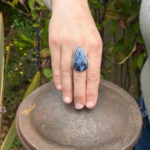 May include: A silver ring with a large, teardrop-shaped stone. The stone is a mix of dark blue and white patterns, set in a simple silver band. The ring is displayed on a hand.