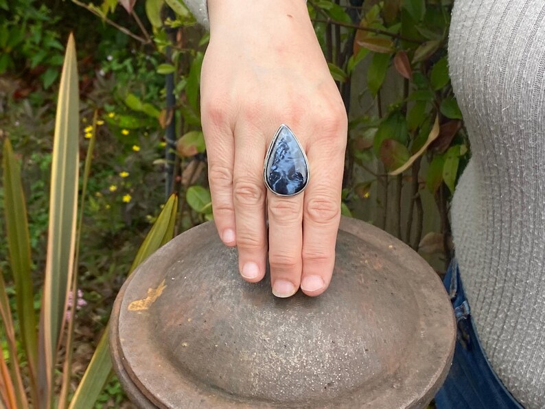 May include: A silver ring with a large, teardrop-shaped stone. The stone is a mix of dark blue and white patterns, set in a simple silver band. The ring is displayed on a hand.