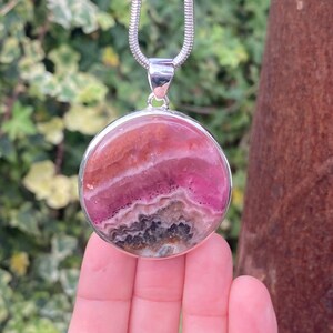 May include: A silver pendant necklace with a round pink and gray gemstone. The gemstone has a layered pattern and is set in a silver bezel.