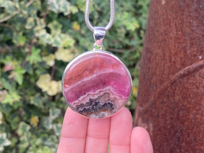 May include: A silver pendant necklace with a round pink and gray gemstone. The gemstone has a layered pattern and is set in a silver bezel.