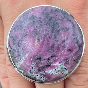 Könnte beinhalten: Ein silberner Ring mit einem großen, runden, rosafarbenen und schwarzen Edelstein. Der Edelstein hat ein wirbelndes Muster und ist in einer silbernen Fassung gefasst.