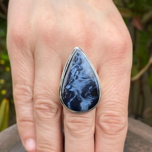 May include: A large, teardrop-shaped ring featuring a polished blue and white stone set in a silver-colored metal band. The stone has a swirling pattern, and the ring is displayed on a hand.