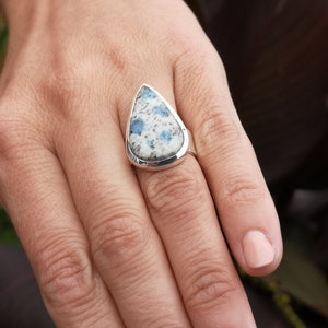 May include: A silver ring with a teardrop-shaped blue and white gemstone. The ring is on a person's finger.