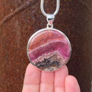 May include: A silver pendant necklace featuring a round, pink and white gemstone with a layered, banded pattern. The pendant is set in a silver bezel.