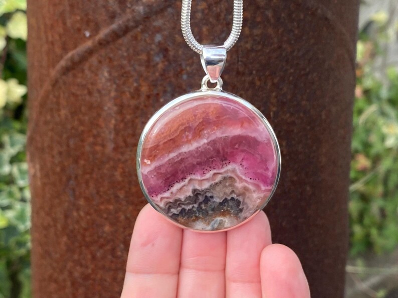 May include: A silver pendant necklace featuring a round, pink and white gemstone with a layered, banded pattern. The pendant is set in a silver bezel.