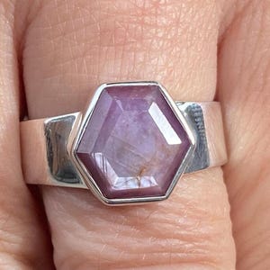 May include: A silver ring featuring a large, hexagonal, light purple gemstone. The gemstone has a faceted surface and is set in a wide, polished silver band. The ring is shown on a finger, highlighting its design and color.