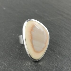 K&ouml;nnte beinhalten: Ein Silberring mit einem hellbraunen und wei&szlig;en Achat-Stein, der in einer Zargenfassung gefasst ist.