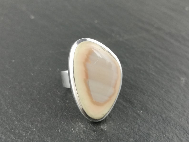 K&ouml;nnte beinhalten: Ein Silberring mit einem hellbraunen und wei&szlig;en Achat-Stein, der in einer Zargenfassung gefasst ist.
