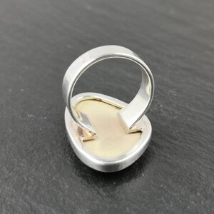 K&ouml;nnte beinhalten: Ein silberner Ring mit einem gro&szlig;en, ovalen, wei&szlig;en Edelstein, der in der Mitte eingesetzt ist. Der Ring wird von hinten gezeigt, wodurch die glatte, silberne Ringschiene sichtbar wird.