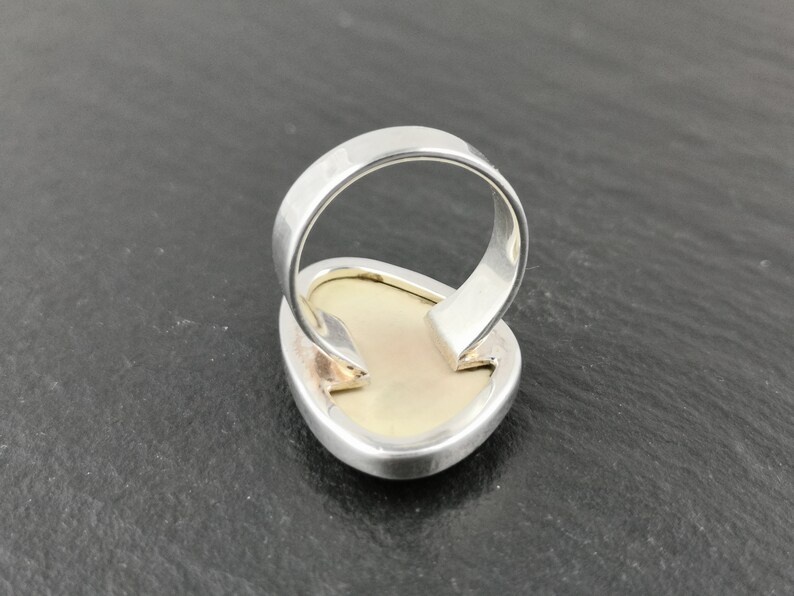 K&ouml;nnte beinhalten: Ein silberner Ring mit einem gro&szlig;en, ovalen, wei&szlig;en Edelstein, der in der Mitte eingesetzt ist. Der Ring wird von hinten gezeigt, wodurch die glatte, silberne Ringschiene sichtbar wird.