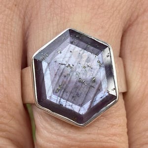 Può includere: Anello in argento con una grande pietra esagonale color lavanda e grigio. La pietra ha un design sfaccettato con inclusioni visibili. L'anello è montato su una fascia larga.