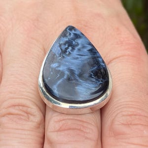 May include: A teardrop-shaped ring featuring a polished blue and black gemstone set in a silver band. The stone has a swirling pattern, creating a unique and eye-catching design. This ring is a statement piece.