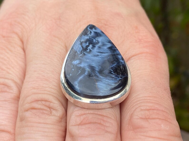 May include: A teardrop-shaped ring featuring a polished blue and black gemstone set in a silver band. The stone has a swirling pattern, creating a unique and eye-catching design. This ring is a statement piece.