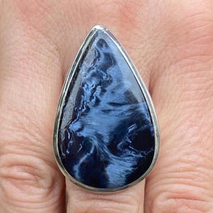 May include: A teardrop-shaped ring featuring a polished blue gemstone with swirling patterns, set in a silver-colored metal band. The gemstone's color ranges from deep navy to lighter shades of blue.