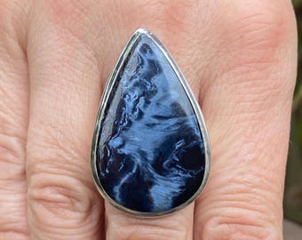 Op de afbeelding: Zilveren ring met een druppelvormige, gepolijste blauwe edelsteen. De steen vertoont wervelende patronen in marineblauw en lichtere blauwtinten. Een sieraad.