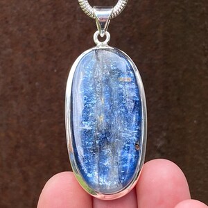 May include: An oval-shaped blue kyanite gemstone pendant set in a polished silver bezel. The stone features deep blue and translucent streaks with a natural crystalline texture. The pendant hangs from a silver snake chain necklace.