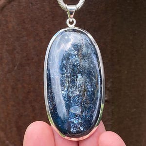 May include: An oval-shaped blue kyanite gemstone pendant set in a polished silver bezel. The stone features deep navy and lighter blue streaks with a reflective, crystalline texture. The pendant hangs from a silver bail attached to a silver chain.
