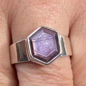 May include: A silver ring featuring a large, hexagonal, purple gemstone. The gemstone has a star-like pattern within it. The ring has a wide, flat band. The ring is shown on a finger.
