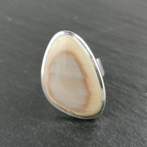 K&ouml;nnte beinhalten: Ein Silberring mit einem hellbraunen und wei&szlig;en Achat-Stein, der in einer glatten, ovalen Form gefasst ist.