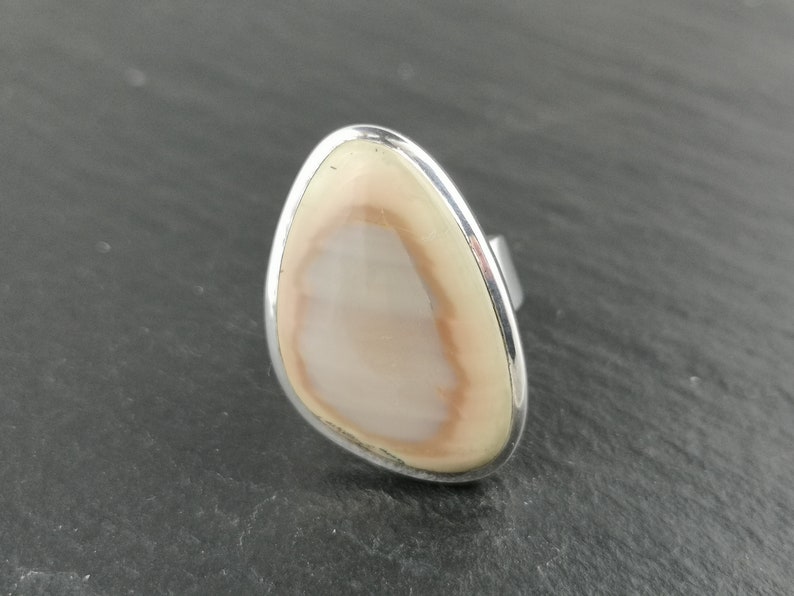 K&ouml;nnte beinhalten: Ein Silberring mit einem hellbraunen und wei&szlig;en Achat-Stein, der in einer glatten, ovalen Form gefasst ist.