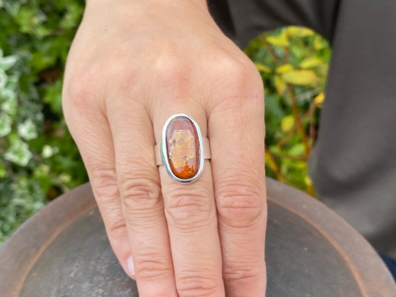 May include: A silver ring with an oval-shaped amber stone. The stone has a translucent, golden-brown color with flecks of white.