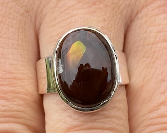 May include: A silver ring with a large, oval-shaped, dark brown gemstone. The gemstone has a reflective surface with hints of yellow and green. The ring band is wide and made of polished silver.