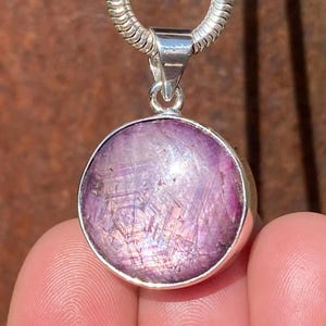 Puede incluir: Un colgante redondo de plata con una gema púrpura grande y pulida. La gema tiene una estructura cristalina interna única. El colgante cuelga de una cadena de plata. El fondo es una superficie marrón borrosa.