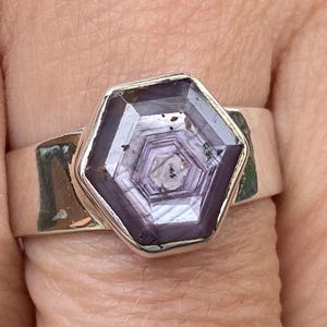 May include: A silver ring featuring a large, hexagonal purple gemstone with a faceted design. The gemstone displays a layered, geometric pattern. The ring is on a finger, showcasing the band's width and the stone's intricate design. A striking piece of jewellery.