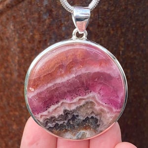 May include: A silver pendant with a round pink and brown gemstone. The gemstone has a layered pattern with shades of pink, brown, and white.