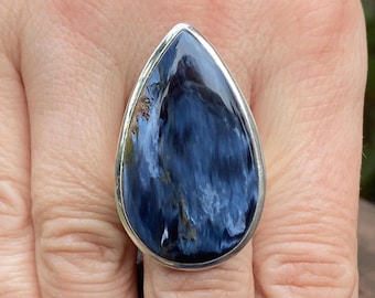 Op de afbeelding: Een opvallende ring met een druppelvormige, gepolijste blauwe edelsteen, gezet in een zilverkleurige bezel. De steen vertoont een mix van donkere en lichte blauwtinten, waardoor een wervelend patroon ontstaat.