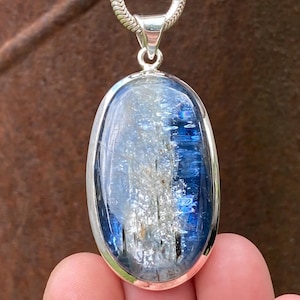 May include: An oval-shaped pendant featuring a polished blue and white kyanite gemstone set in a smooth silver bezel. The stone displays deep blue streaks and translucent white inclusions. It hangs from a silver chain with a braided texture.