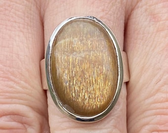 May include: A silver ring featuring an oval-shaped, golden-brown gemstone. The stone has a shimmering, textured appearance, set in a polished silver band. The ring is shown on a finger, highlighting the jewelry's design and color.