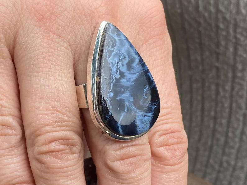 May include: A large, teardrop-shaped ring featuring a polished blue and white gemstone set in a silver band. The stone has a swirling pattern, and the ring is displayed on a finger.