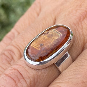 May include: A silver ring with an oval-shaped amber stone set in a bezel setting. The stone has a brown and gold color with a swirling pattern.