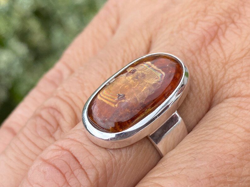 May include: A silver ring with an oval-shaped amber stone set in a bezel setting. The stone has a brown and gold color with a swirling pattern.