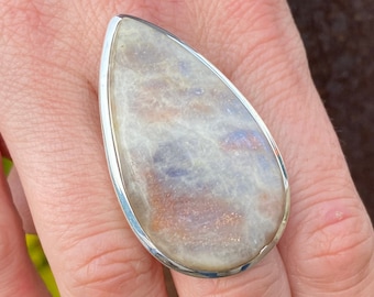 Könnte beinhalten: Ein silberner Ring mit einem großen, tropfenförmigen Mondstein. Der Stein hat einen weißen, irisierenden Glanz mit einem Hauch von Rosa und Blau.