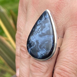 May include: A large, teardrop-shaped ring featuring a polished, dark blue gemstone with swirling white patterns. The stone is set in a silver-colored metal band. The ring is a statement piece of jewelry.