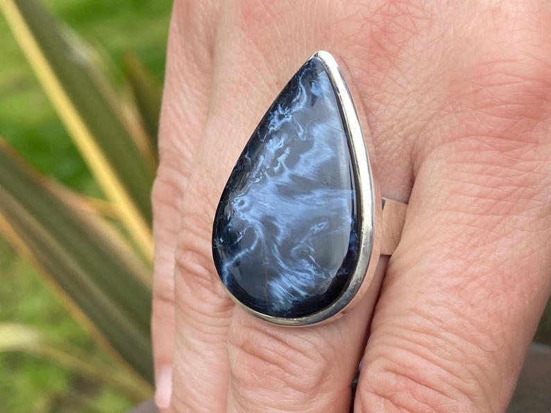 May include: A large, teardrop-shaped ring featuring a polished, dark blue gemstone with swirling white patterns. The stone is set in a silver-colored metal band. The ring is a statement piece of jewelry.
