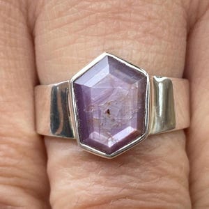 May include: A silver ring featuring a large, hexagonal, lavender-colored gemstone. The gemstone is set in a bezel setting, and the band is wide and polished. The ring is shown on a finger.